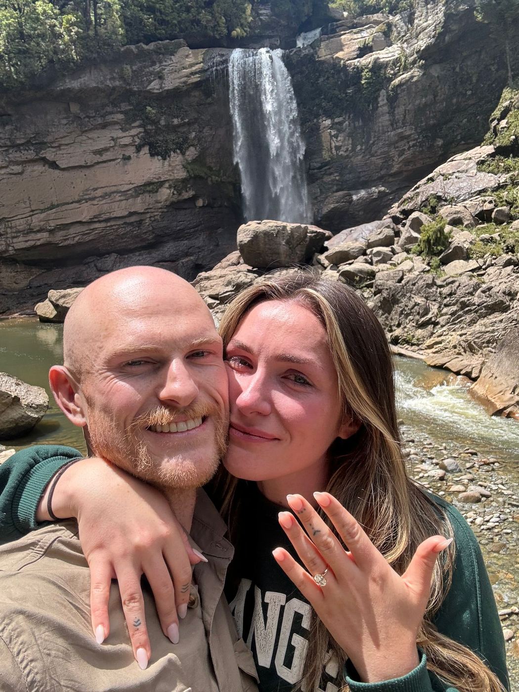 Reece and Robyn moments after their proposal, in front of a waterfall — Robyn showing her engagement ring